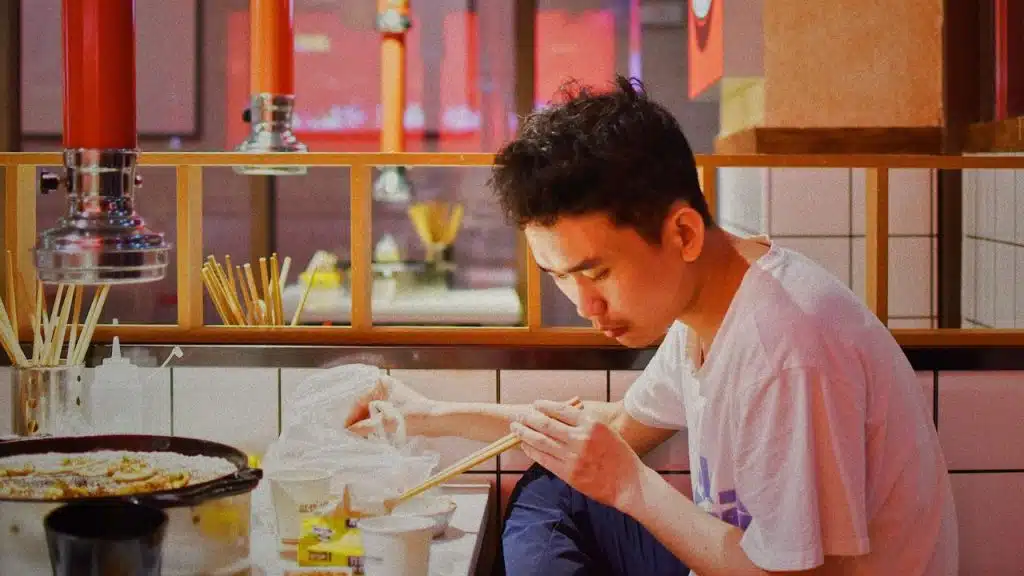 A man in a white t-shirt uses chopsticks to eat at a restaurant table.