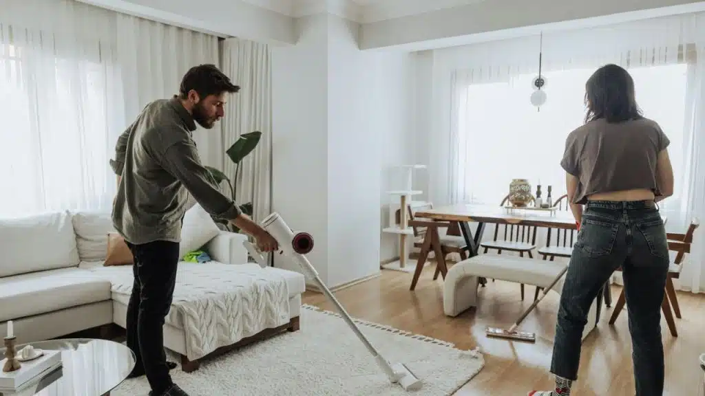 A man vacuums a white rug while a woman stands nearby in a bright room.