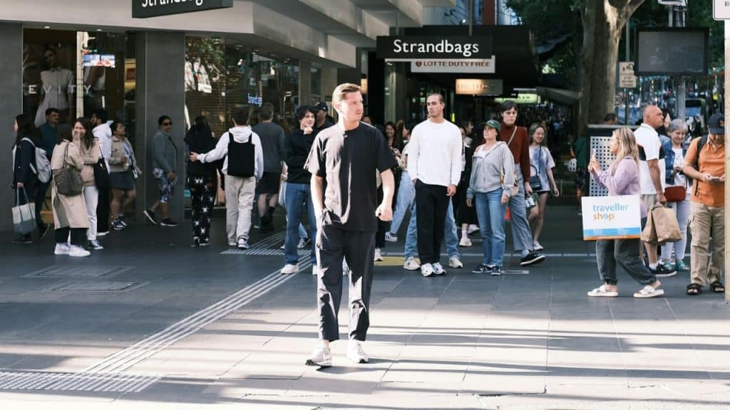 A man in black clothing walks through a crowded city sidewalk in front of storefronts.