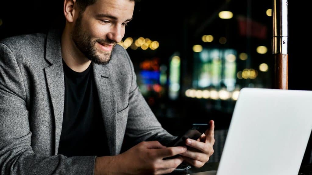 A man smiling while using a phone beside a laptop in a café.