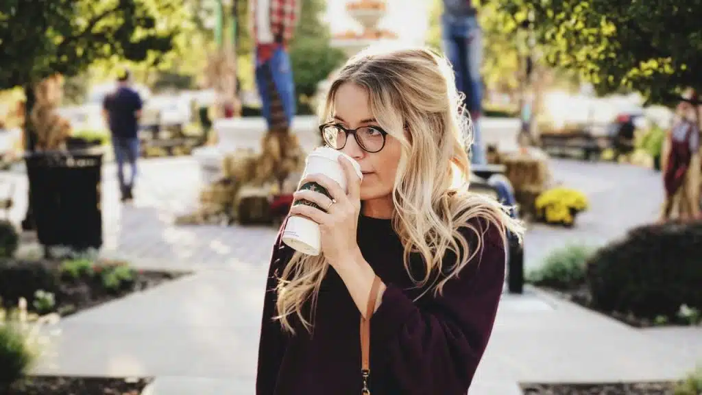 A woman wearing glasses drinking coffee while walking outdoors.