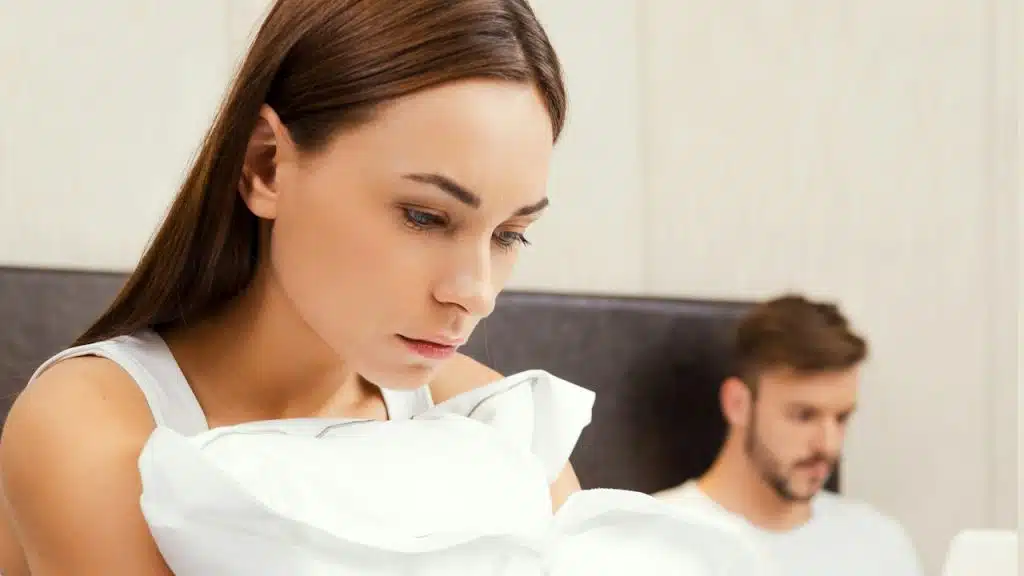 A woman holding a pillow while she is sitting on the bed with her angry husband.