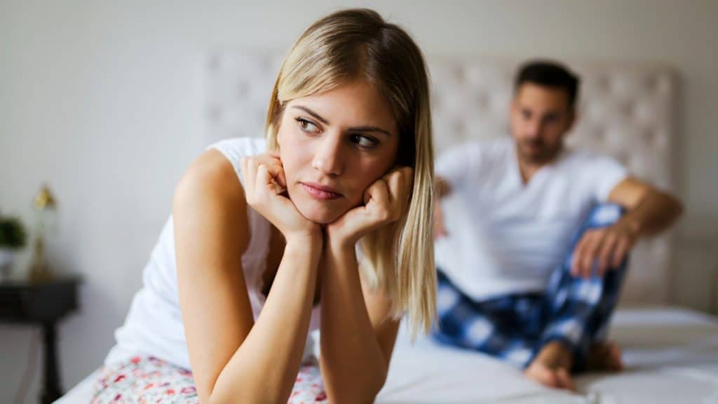 A woman sitting on a bed’s edge and holding her head in her hands while a man sits on the bed behind her.