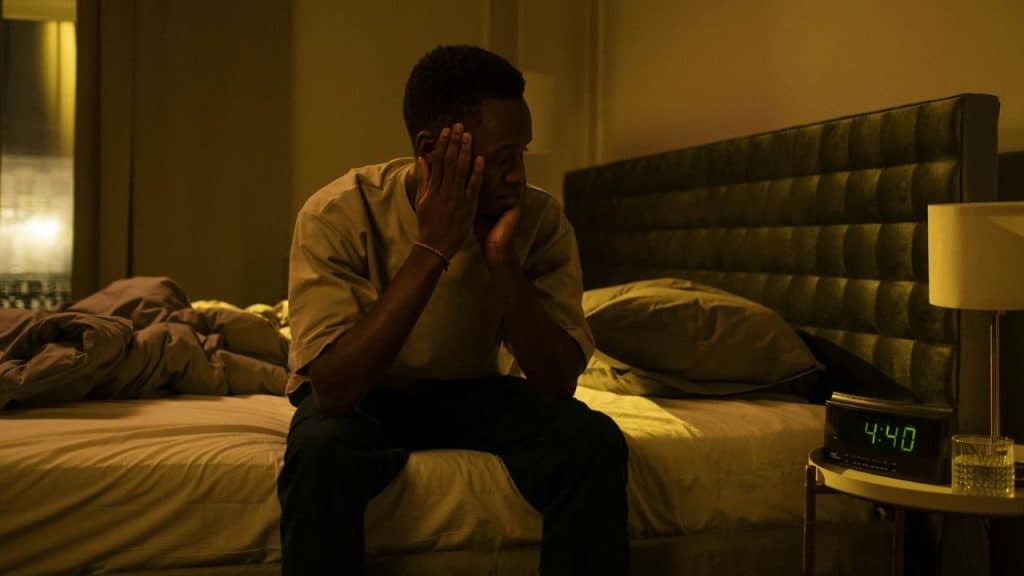 A man sits on a bed holding his head next to a digital clock reading 4:40.
