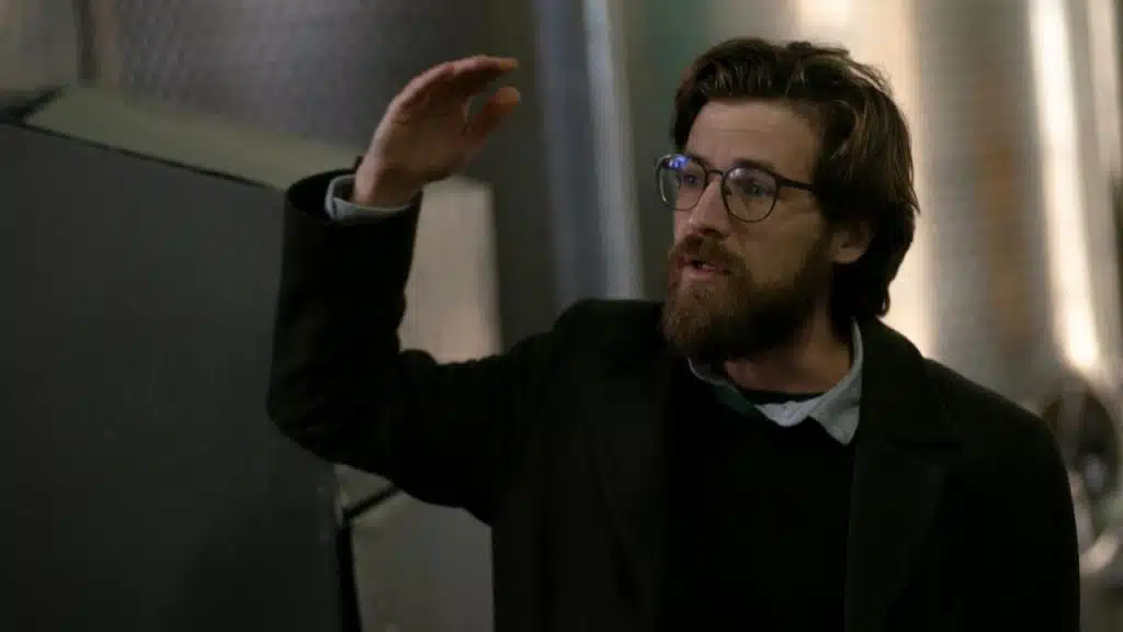 A bearded man wearing glasses and a dark coat gestures while speaking in a room.