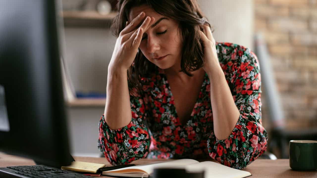 A woman in a floral shirt sits at a desk with her hand on her forehead.