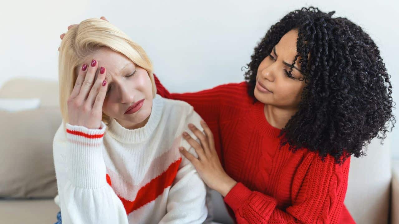 A woman in a red sweater comforts a blonde woman who has her eyes closed.