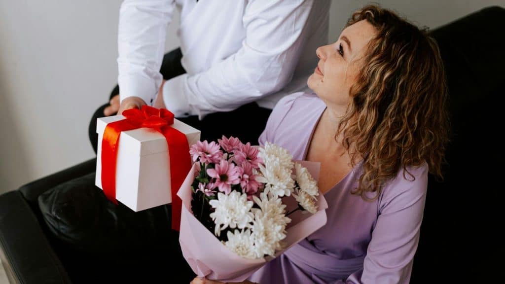 A woman holding flowers looks up at a person holding a white gift with red ribbon.