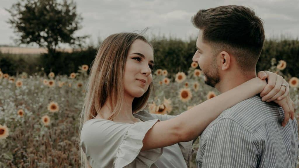 A woman rests her arm on a man’s shoulder while standing in a sunflower field.