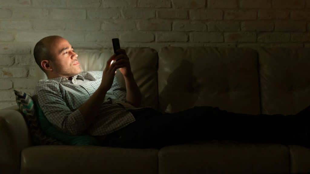 A man lies on a leather couch in a dim room while looking at a smartphone.
