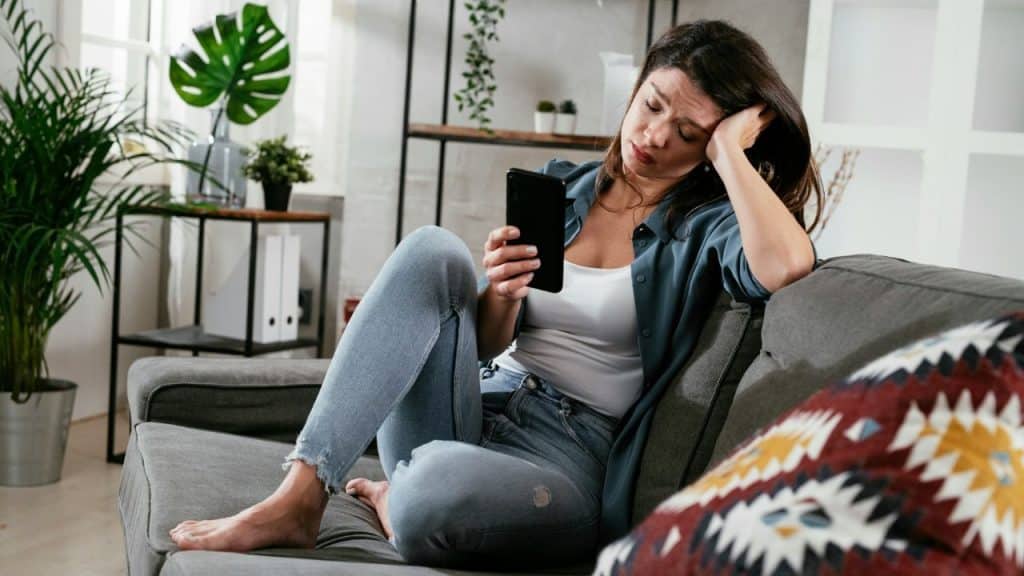 A woman sits on a grey sofa looking down at a phone in her hand.