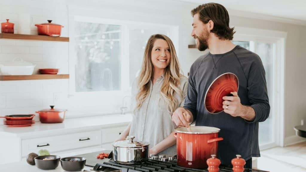 man stirs a red pot while a woman watches him in a bright white kitchen.