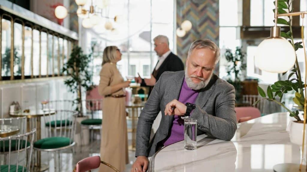 A man in a grey blazer checks his watch while leaning on a marble bar counter.