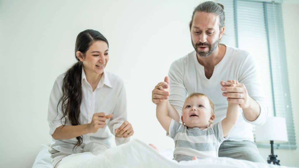 A man and woman play with a baby on a white bed in a bright room.