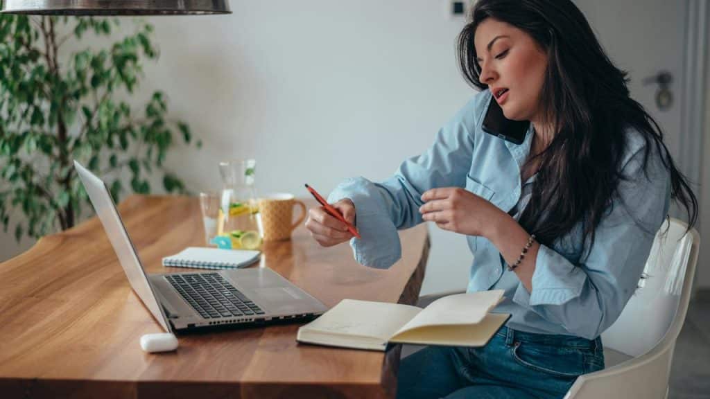 A woman multitasking with a phone on her shoulder while working on a laptop.