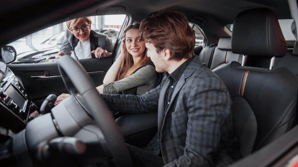 A car salesperson speaking with a couple seated inside a vehicle.