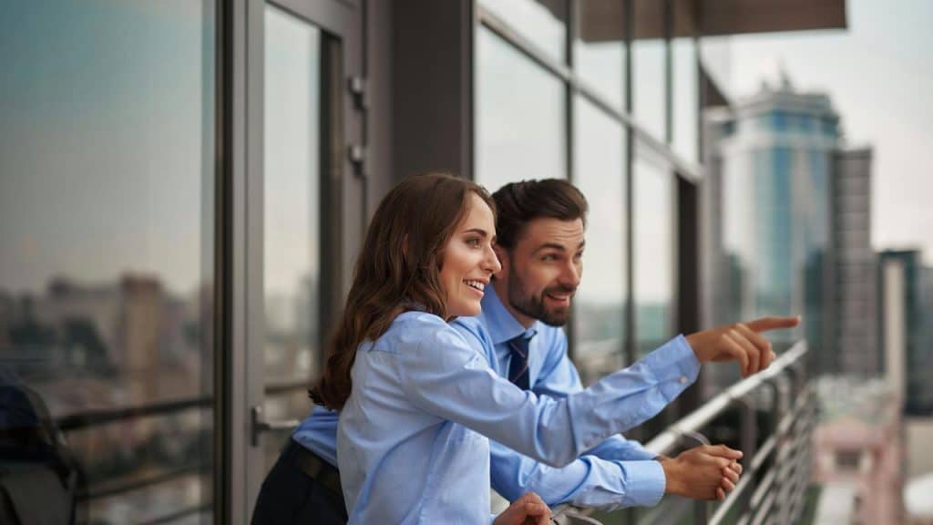 A man and a woman in business attire smiling while looking out from a balcony.