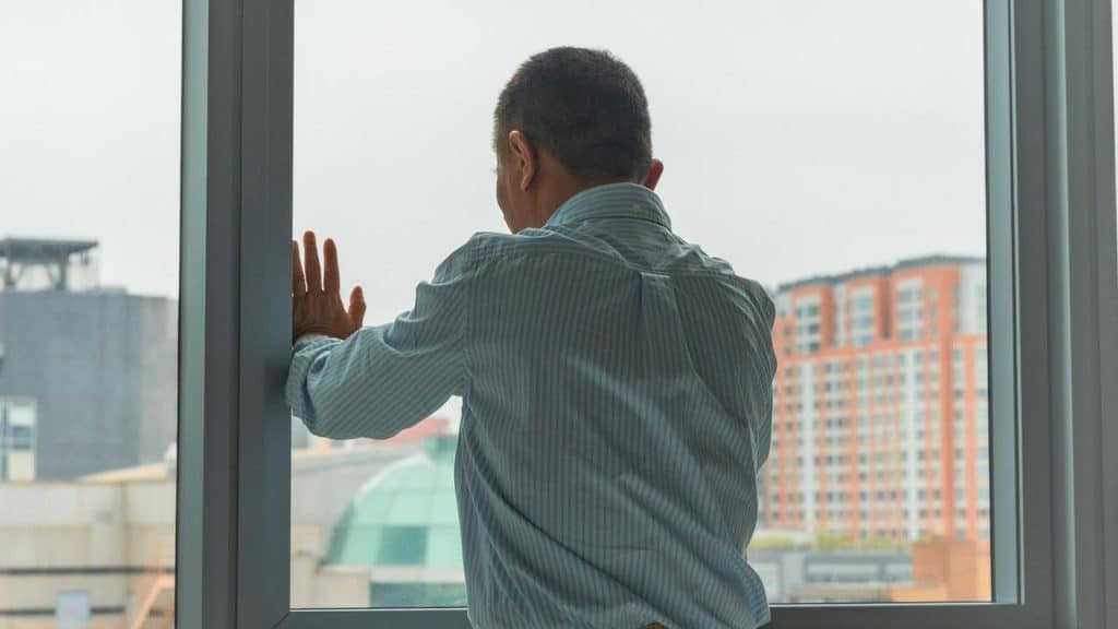 A man in a striped shirt standing by a window with his hand on the glass.