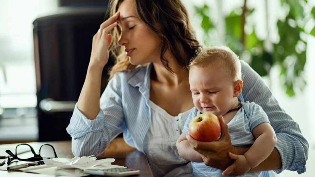 A tired mother holds a crying baby with an apple.