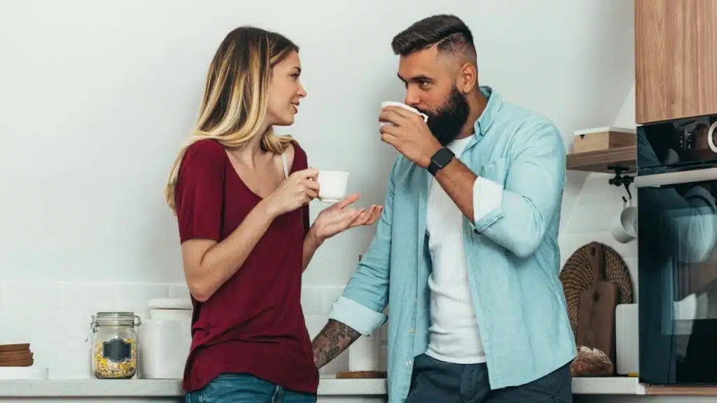A man and woman stand in a kitchen holding white mugs and talking together.
