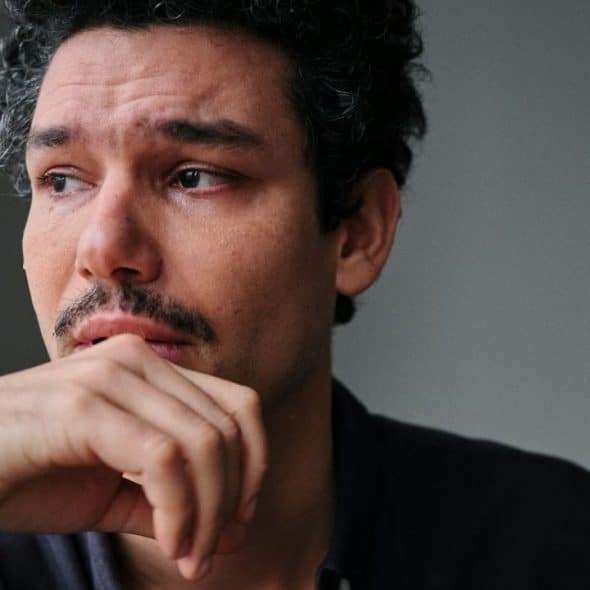 A man with dark curly hair and a mustache looking away with a tearful expression.
