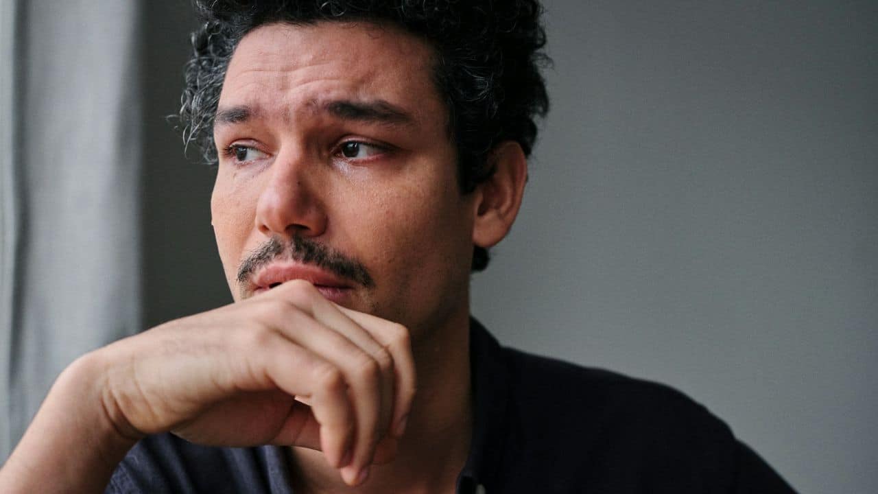 A man with dark curly hair and a mustache looking away with a tearful expression.