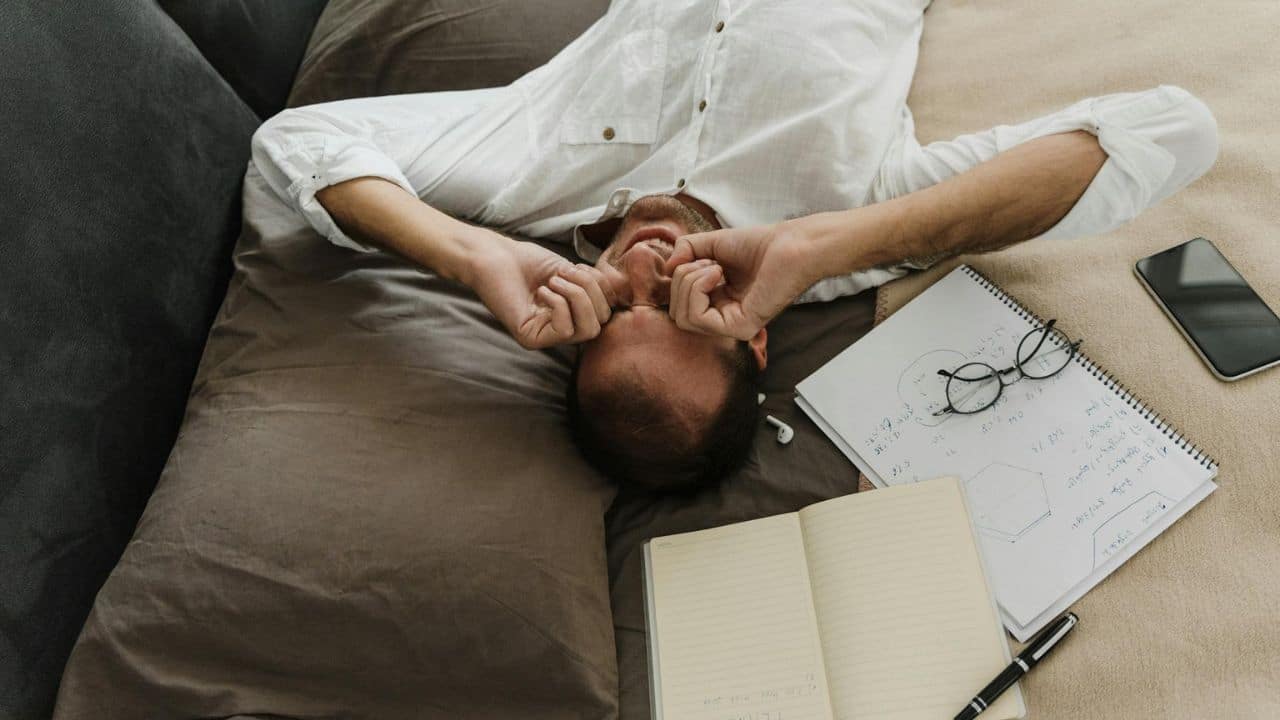 A man lies on a bed rubbing his eyes next to open notebooks and pens.