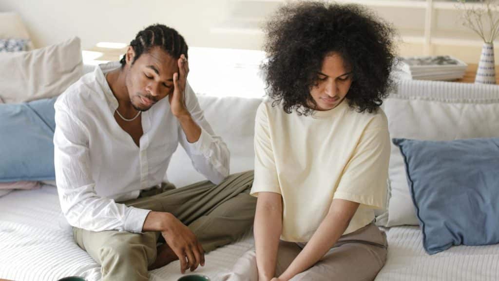 A man and woman sit apart on a light-colored sofa looking down with somber expressions.