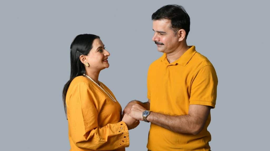 A man and woman in yellow clothing stand facing each other, holding hands and smiling.
