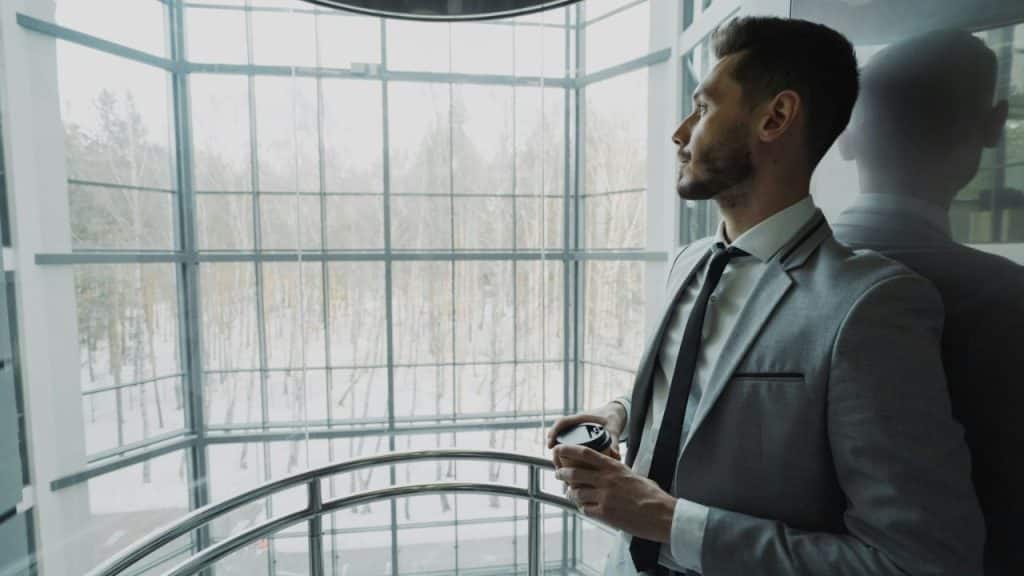 A man in a light grey suit looks out a glass elevator window while holding coffee.