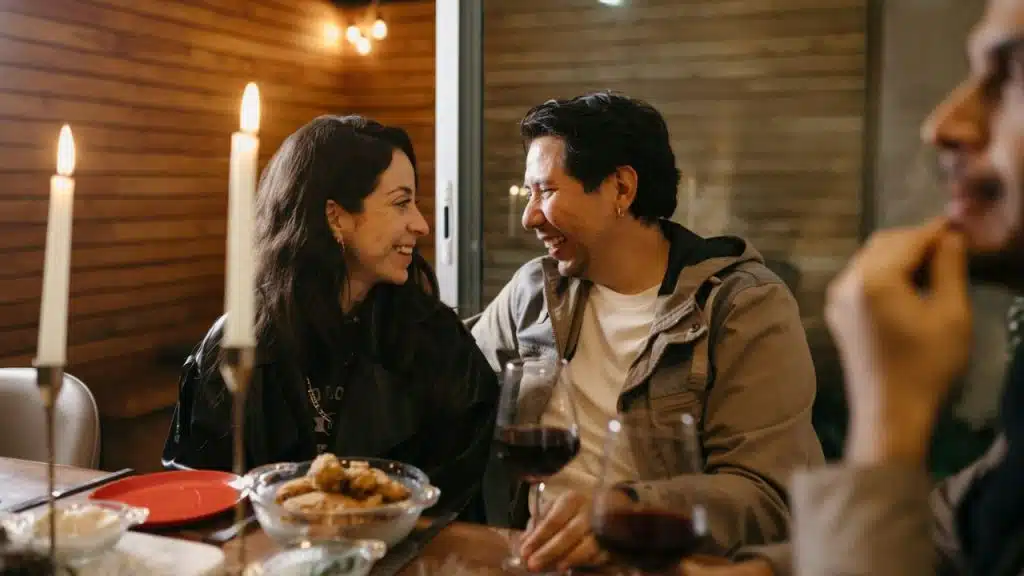 A woman in a black jacket and a man in a tan jacket laugh together at dinner.
