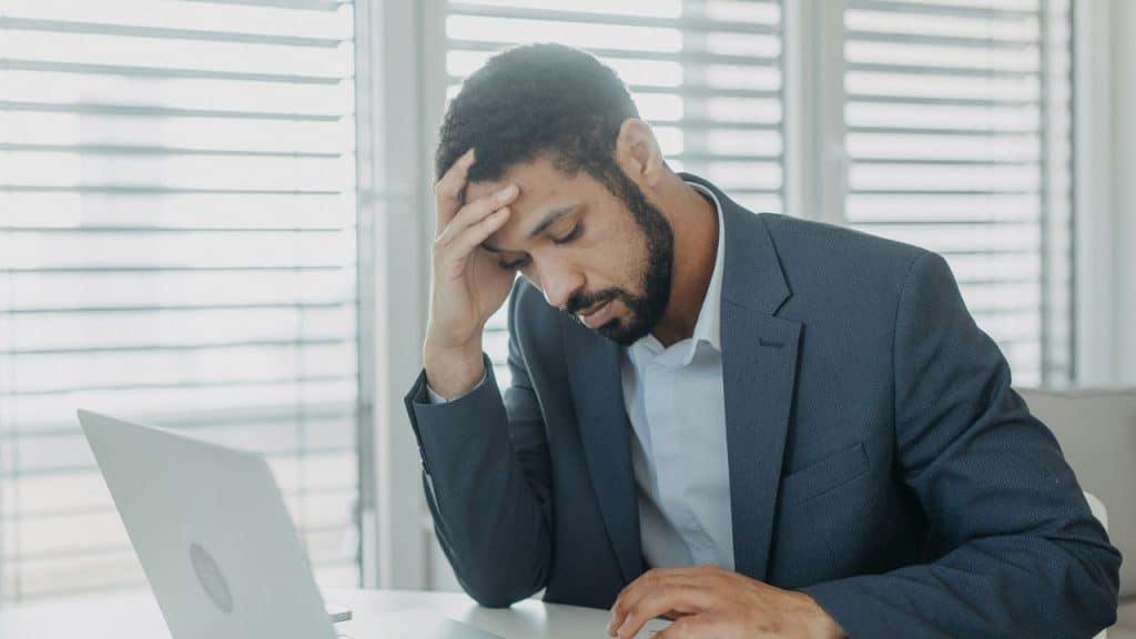 A man in a suit sits at a desk with his hand resting on his forehead.