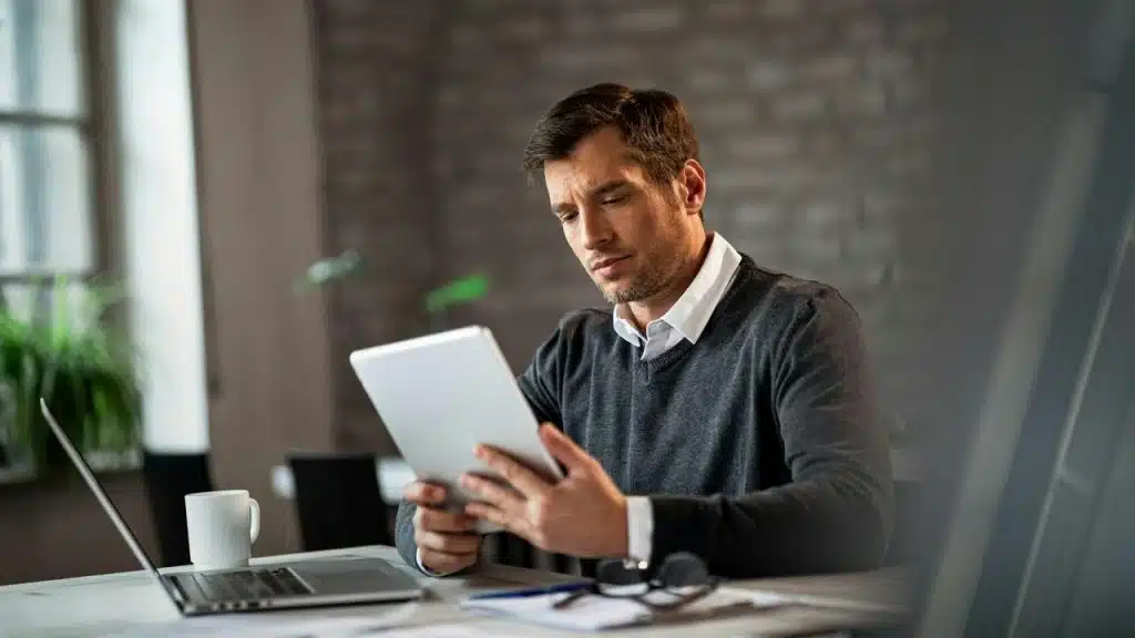 A man sitting at a desk using a tablet beside a laptop.