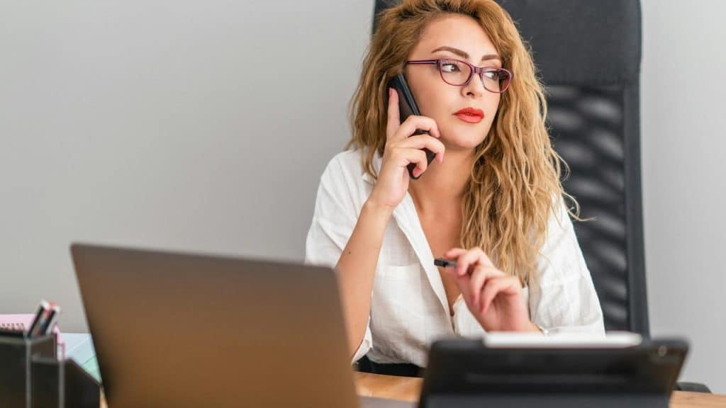 A woman wearing glasses talking on a phone while sitting at a desk with a laptop
