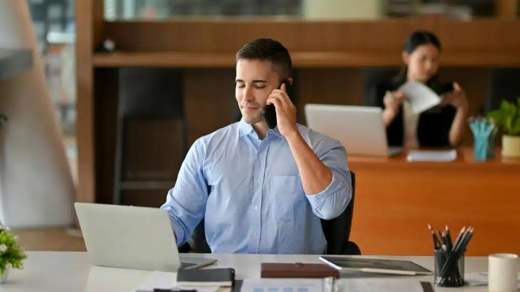 A man in a blue shirt talking on a phone while working on a laptop at a desk.