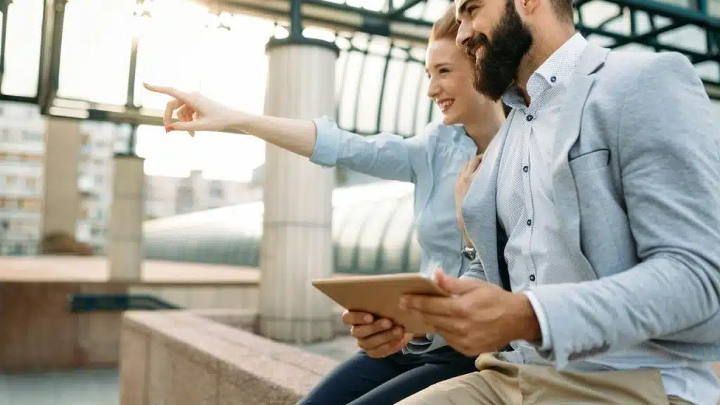 A smiling woman pointing while sitting beside a man holding a tablet.
