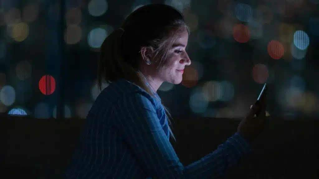 A young woman smiling at her phone at night with blurred city lights in the background.