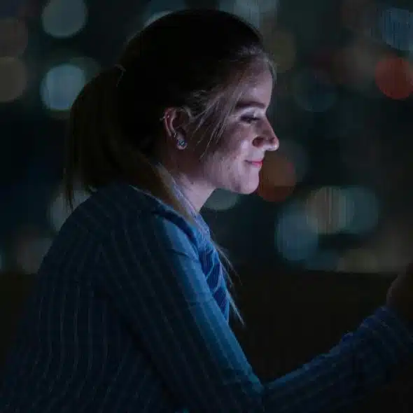 A young woman smiling at her phone at night with blurred city lights in the background.