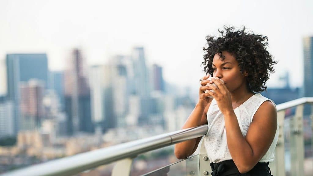 A woman drinking from a cup on a balcony overlooking a city skyline.