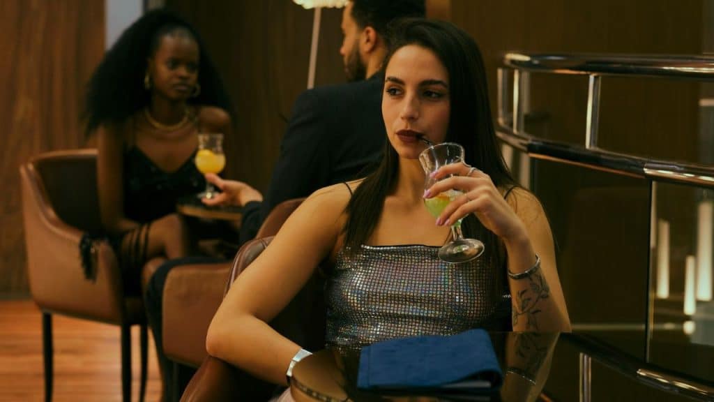 A woman in a sparkly silver top drinks a green beverage through a black straw.