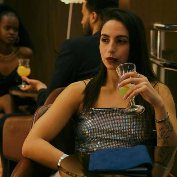 A woman in a sparkly silver top drinks a green beverage through a black straw.