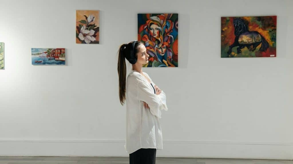 A woman with a ponytail and headphones looks at colorful paintings in a white gallery.