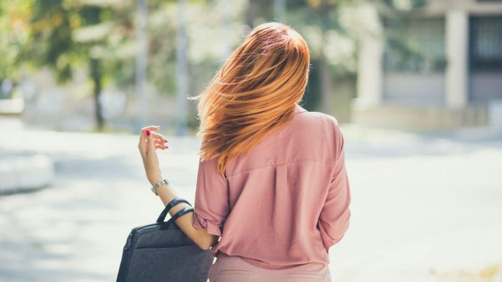 A woman with long reddish-orange hair walks away outdoors while carrying a grey laptop bag.
