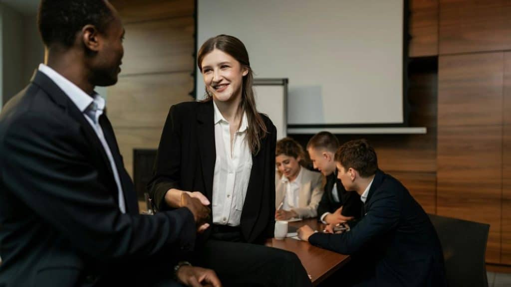 A woman smiles and shakes hands with a man while others sit at a table.