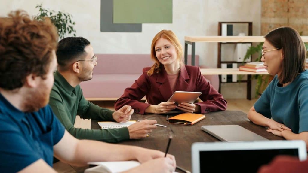 Four people sit around a wooden table while a woman in a maroon blazer talks.