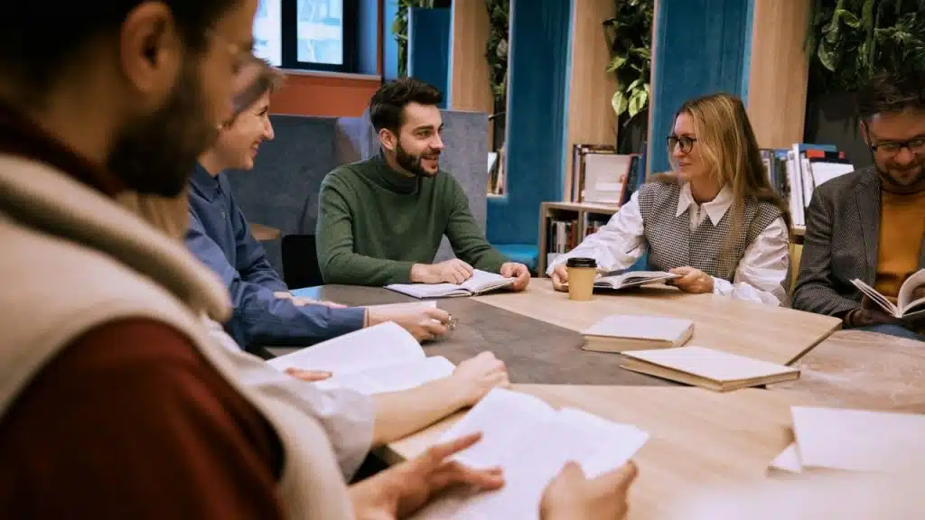 Five people sit around a table with books and coffee in a modern office space.