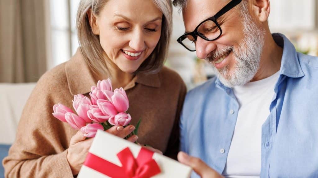 A smiling woman holding pink tulips and a man in glasses looking at a gift.
