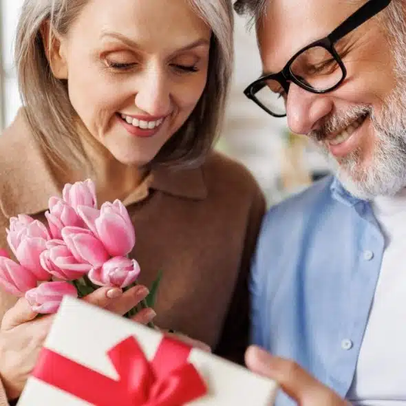 A smiling woman holding pink tulips and a man in glasses looking at a gift.