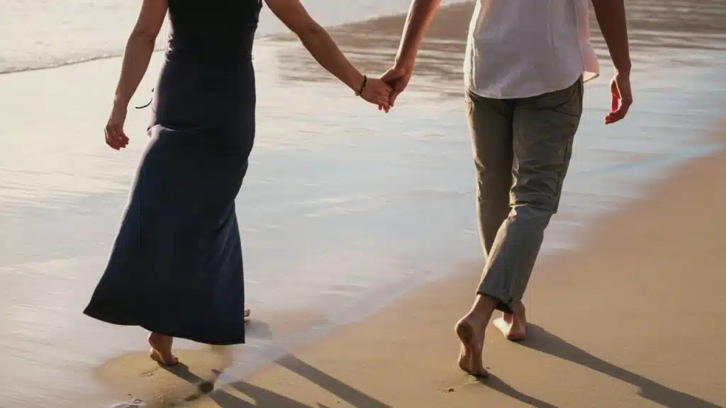 A man and a woman walk barefoot on a sandy beach while holding each other's hands.