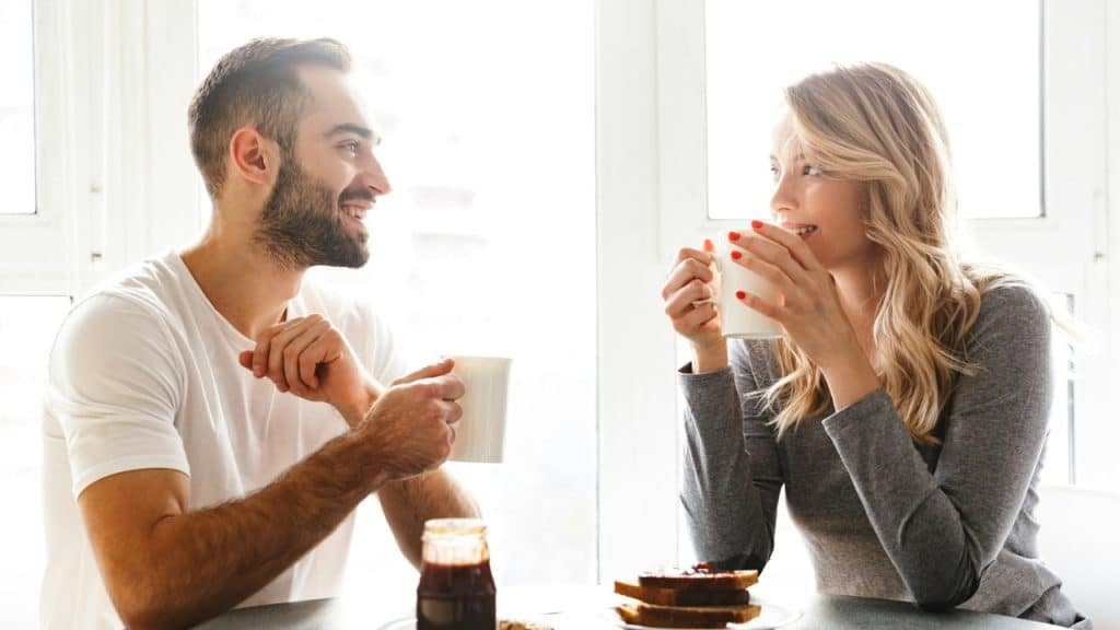 A man and woman sit at a table drinking from white mugs near toast and jam.